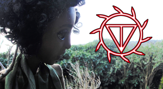 Image shows a puppet of a pensive, dark-skinned child in front of a wild hedge 
and a blank sky emblazoned with a mysterious red icon.