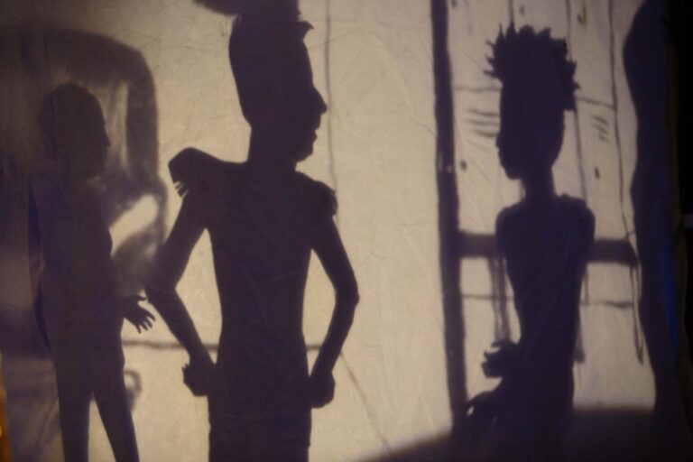 Shadow puppets of Clay Odinson, a tall man with a domineering pose, and Peat Orson, a young man with an Afro, cut short at the sides.