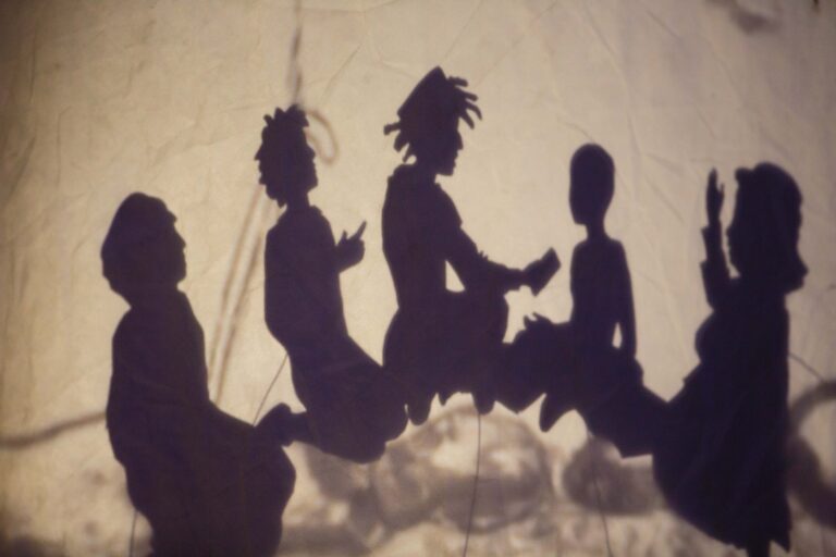 A shadow puppetry scene of a family sitting in a tent. There is a white crook-handled cane in the background.