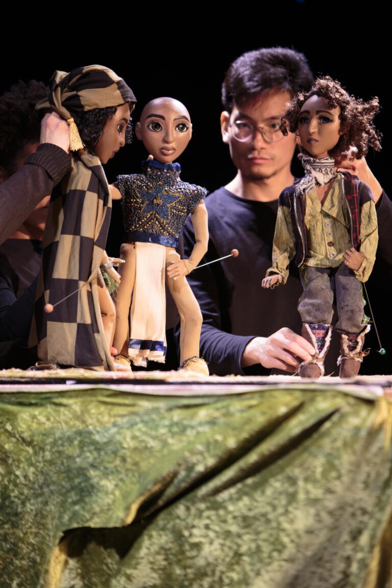 Puppets Lilt, in a hooded cloak, Astra, with shaven head, and Muzi the shepherd, held by Raven Kaliana, Sara Guedes and Gun Suen